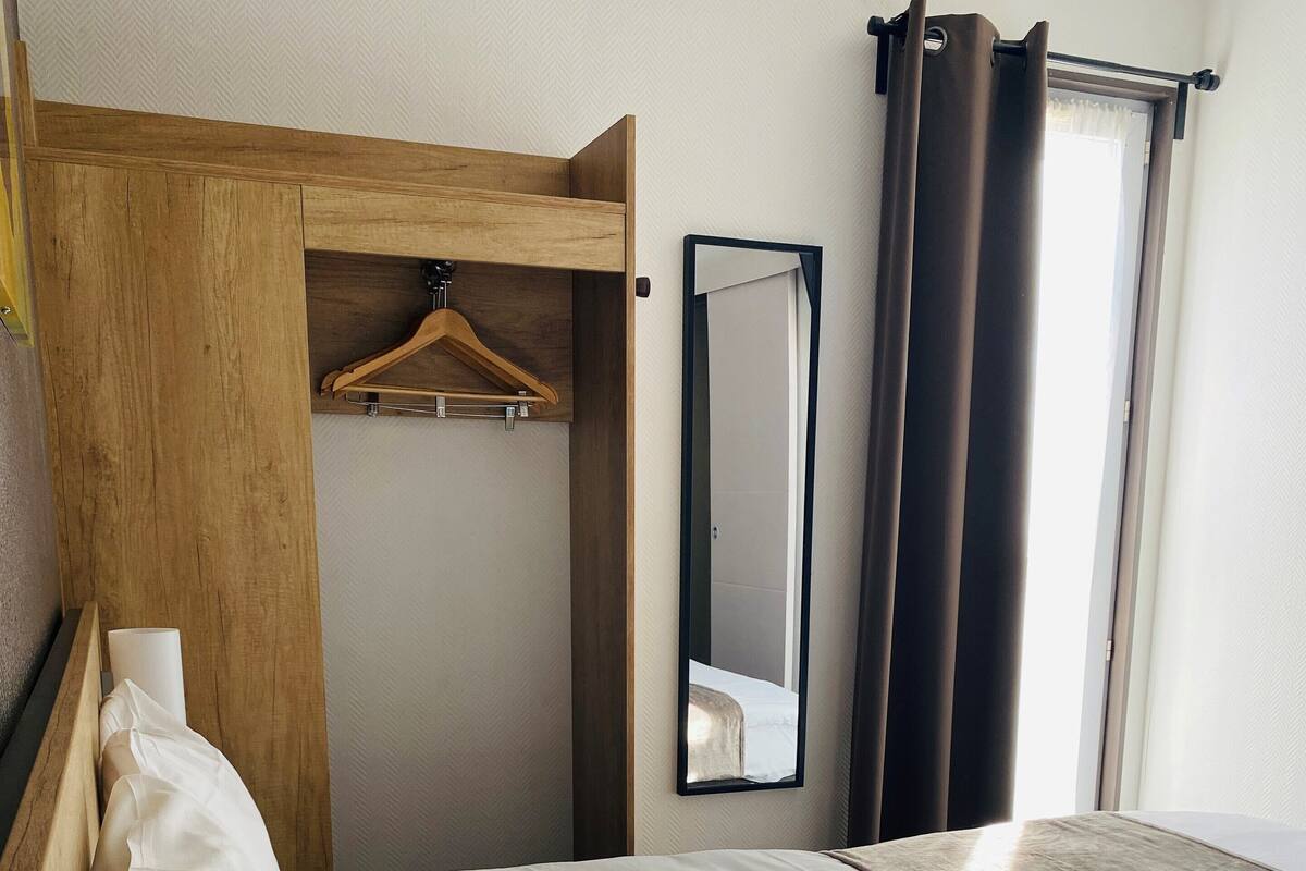 Een hotelkamer met een net opgemaakt bed, een houten garderobe, een spiegel en een raam met gordijnen.