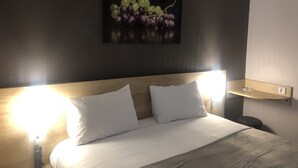 Hotelkamer met een bed, witte kussens, een nachtkastje met een afstandsbediening, een wandlamp en een schilderij van druiven.