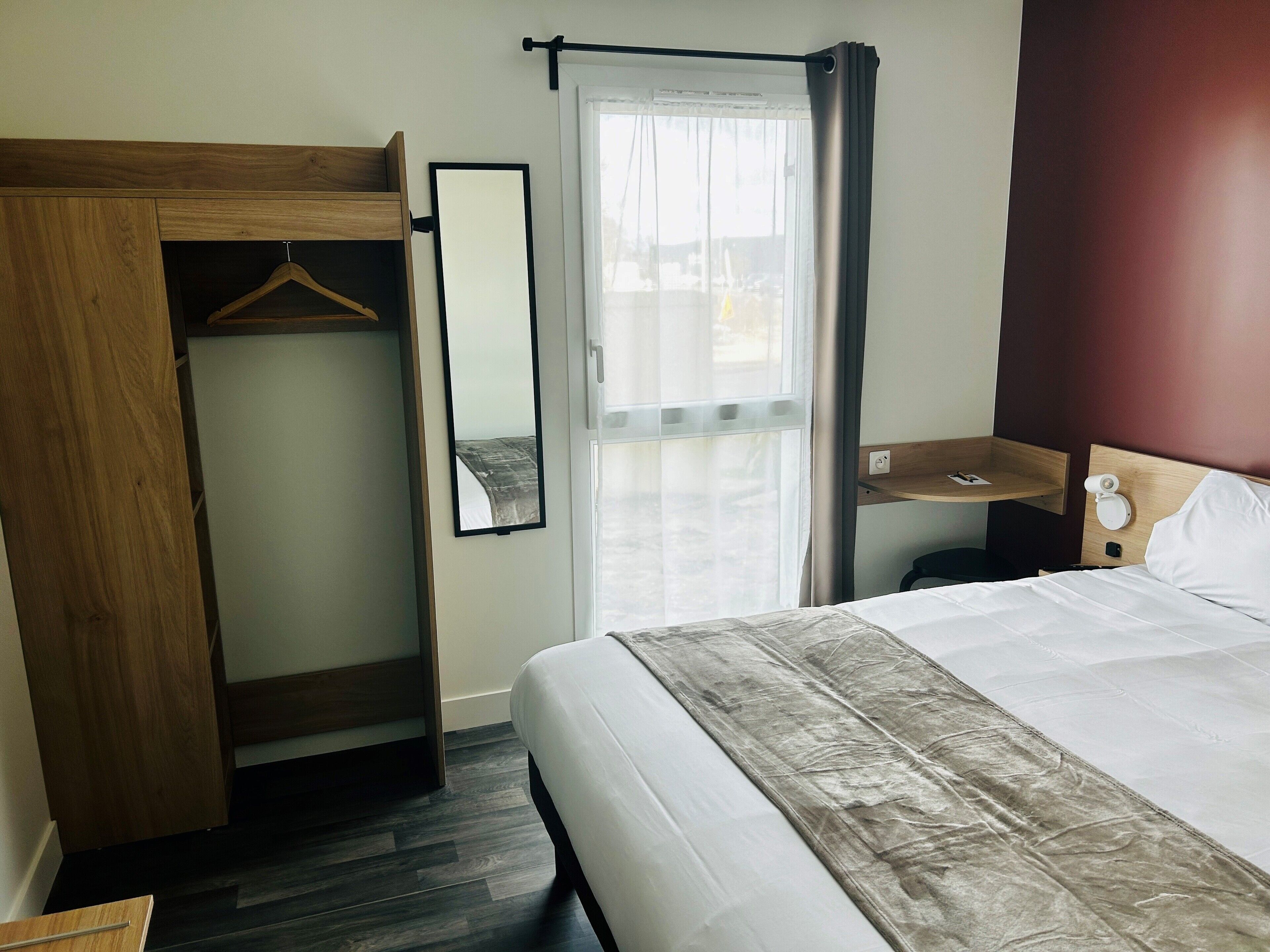 A hotel room with a bed, a wooden wardrobe, a mirror, a bedside table with a lamp, and a window with curtains.