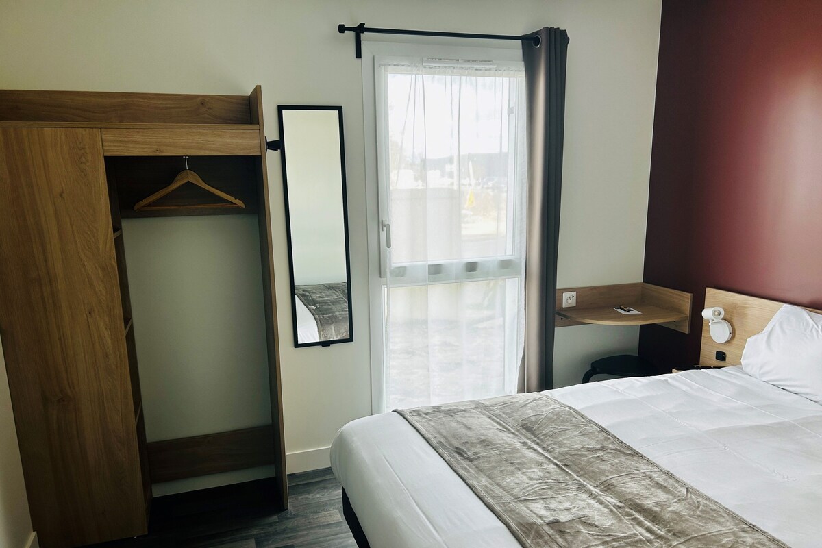 Ein Hotelzimmer mit Bett, Holzschrank, Spiegel, Nachttisch mit Lampe und Fenster mit Vorhängen.