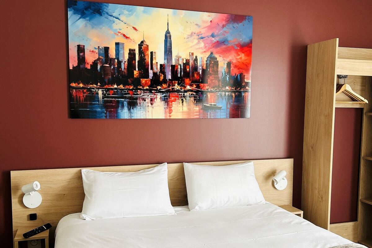 Een hotelkamer met een bed, nachtkastje, houten hoofdeinde, schilderij van een stadshemelbeeld en een houten garderobe.
