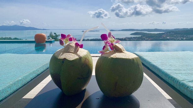 Deux noix de coco, chacune munie d’une paille et décorée de fleurs, sont posées sur une table au bord de la piscine, d’où l’on peut admirer la piscine et un paysage magnifique avec des montagnes et de l’eau.