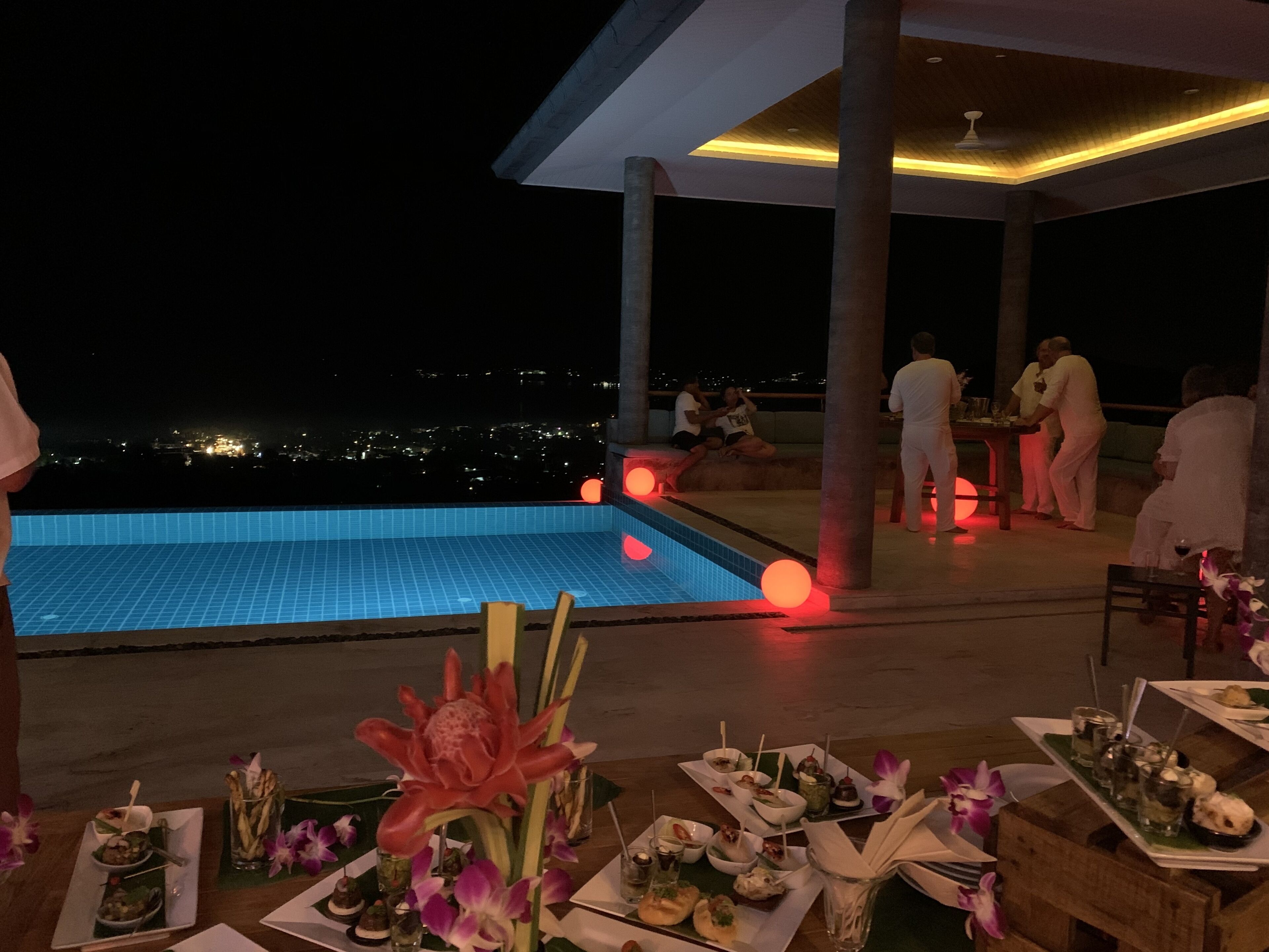 A rooftop dining area with a pool, tables set for a meal, and guests enjoying the evening.