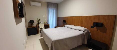 A hotel room with a bed, a desk, a chair, a TV, and an air conditioning unit.