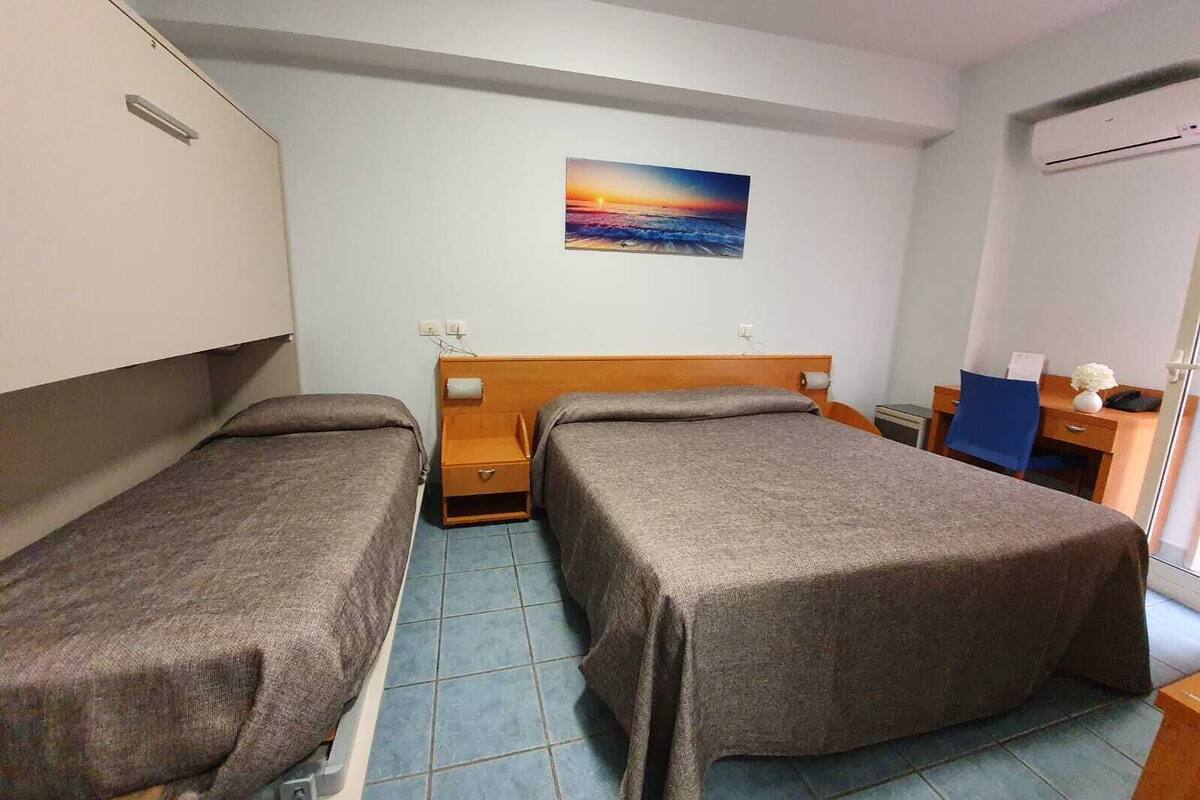 A hotel room with two single beds, a wooden nightstand, a desk with a chair, a painting on the wall, and an air conditioning unit.