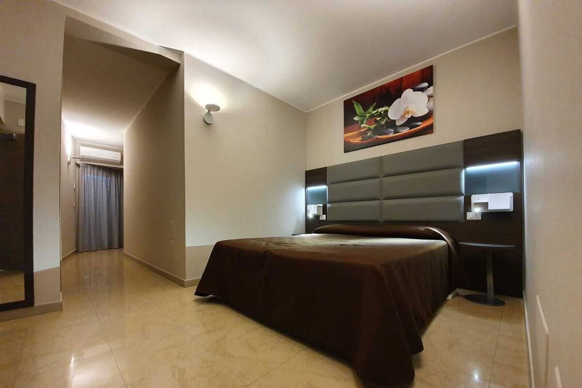 A modern hotel room with a large bed, a headboard with built-in lighting, a painting of flowers on the wall, and a small desk with a chair.