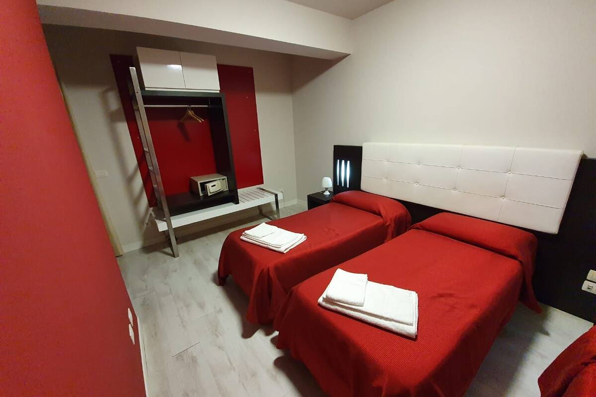 A hotel room with two beds, a red wall, a wardrobe, and a small table with a lamp.