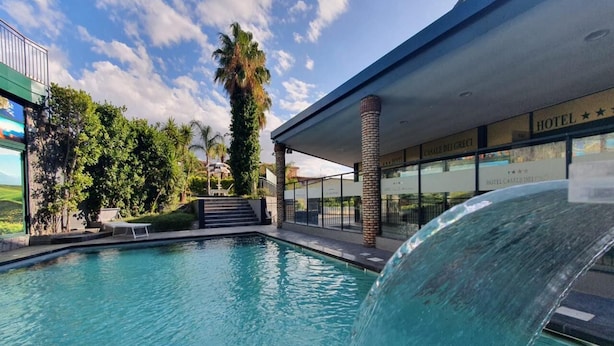 A swimming pool with a waterfall feature, surrounded by a modern building with glass windows and a palm tree. - Hotel Casale dei Greci (Biancavilla)