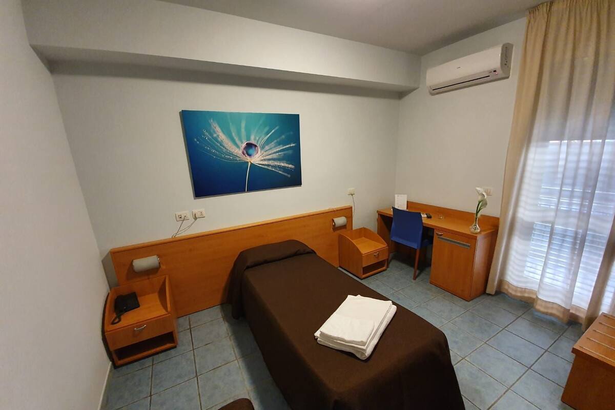 A hotel room with a bed, a desk, a chair, a nightstand, a small table, a vase with a flower, an air conditioner, and a painting on the wall.