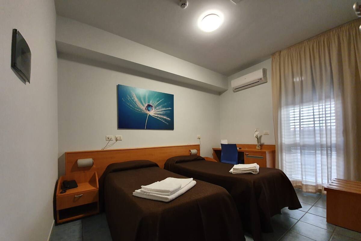 A hotel room with two beds, a desk, a chair, a painting, and an air conditioning unit.