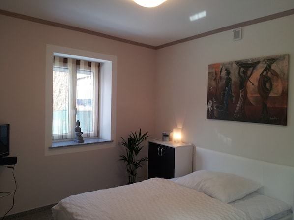 Un dormitorio con cama, ventana con cortinas, un cuadro en la pared y un mueble pequeño con lámpara.