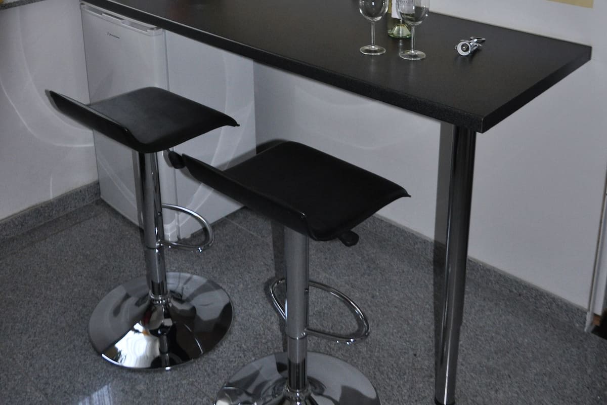A compact kitchen with a black countertop, two bar stools, a microwave, and a small shelf with a plant and a bowl of fruit.