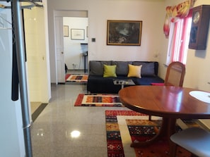 Una sala de estar con sofá, mesa de comedor y una silla. Hay un cuadro en la pared y un reloj.