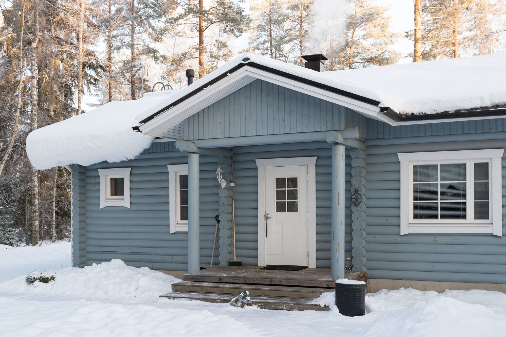 A light blue wooden cabin with a white door and windows, surrounded by snow and trees.