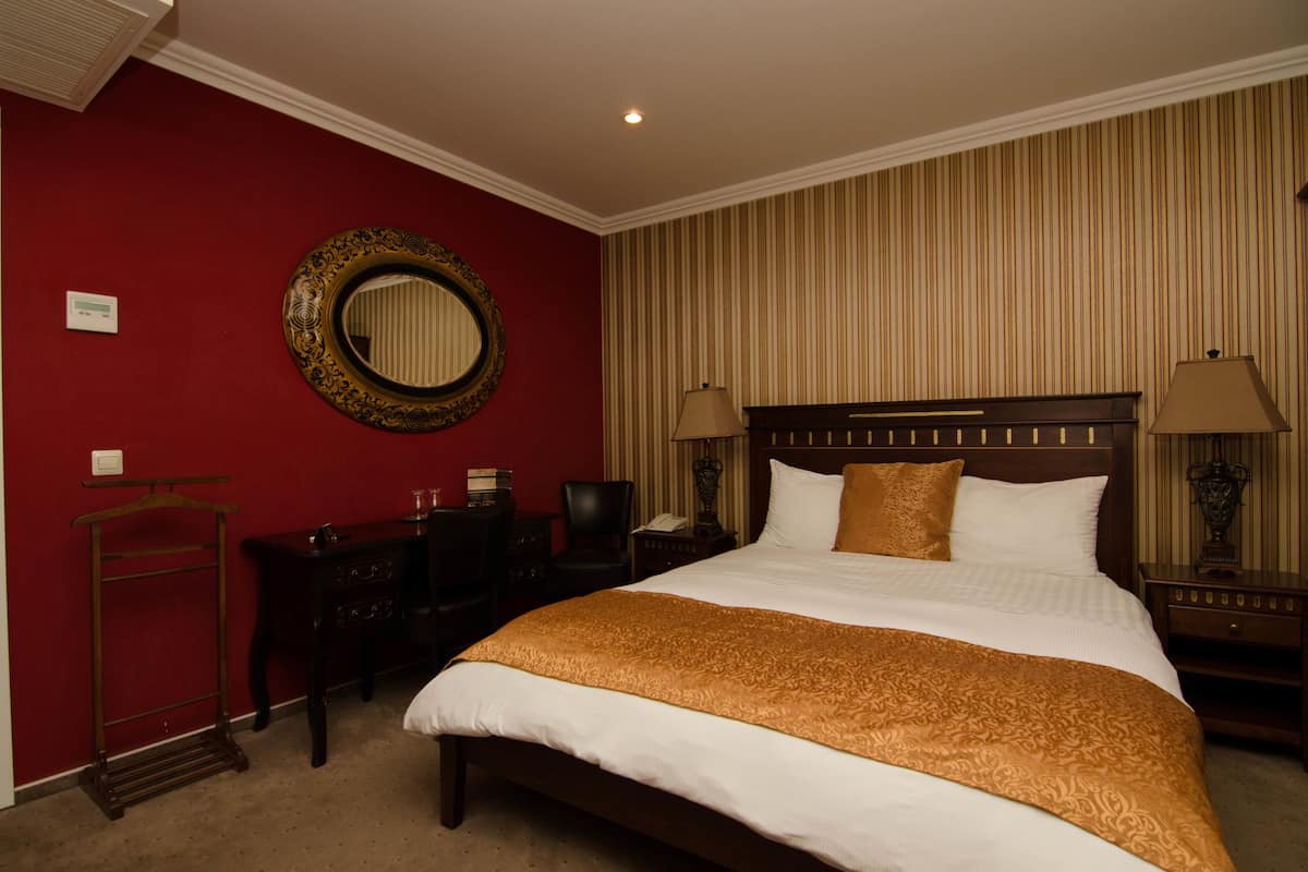 A hotel room with a large bed, a wooden nightstand, a desk with a chair, a mirror, and striped wallpaper.