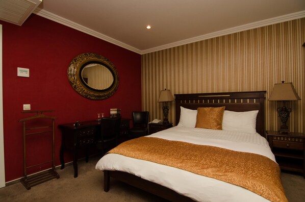 Quarto de hotel com uma cama grande, uma mesa de cabeceira de madeira, uma escrivaninha com cadeira, um espelho e papel de parede listrado.