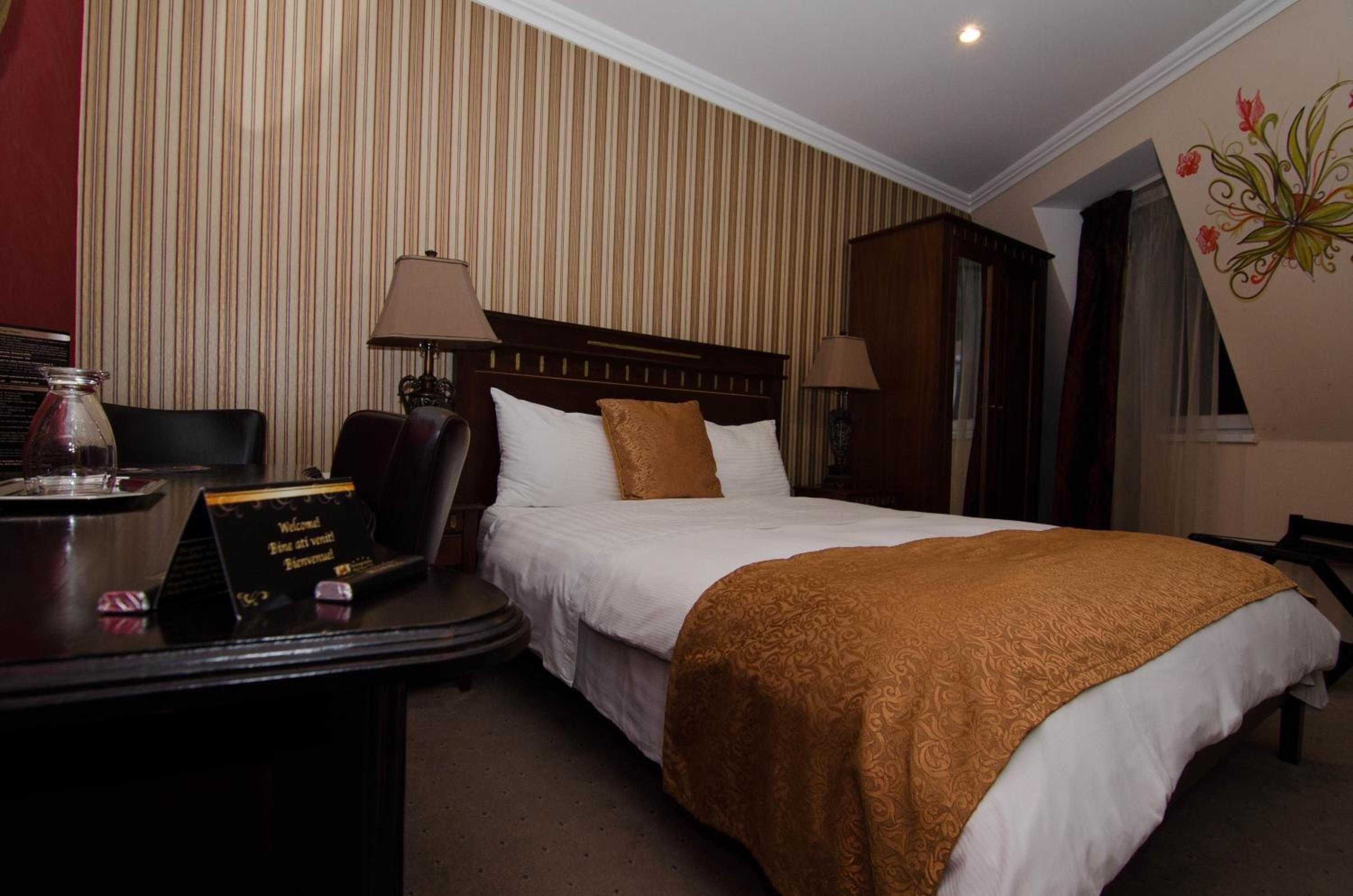 Habitación de hotel con cama, dos lámparas de noche, un escritorio con cartel de bienvenida, una silla y un panel decorativo en la pared.