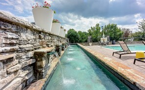 Una piscina bordeada de piedra con una fuente de agua azul cristalina, rodeada de sillones de descanso y plantas en macetas.