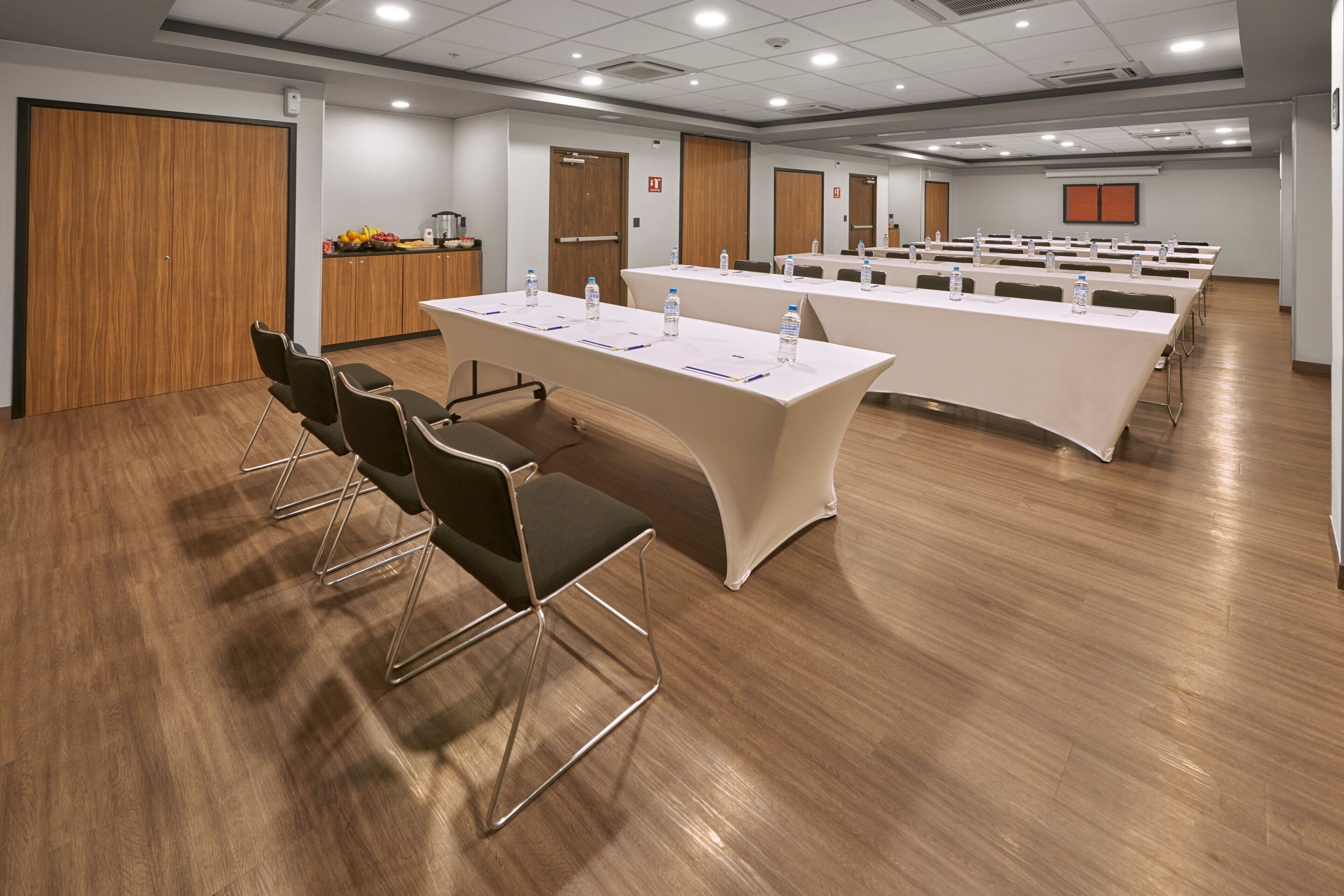 Un salón de conferencias con mesas largas, sillas y una zona de kitchenette.
