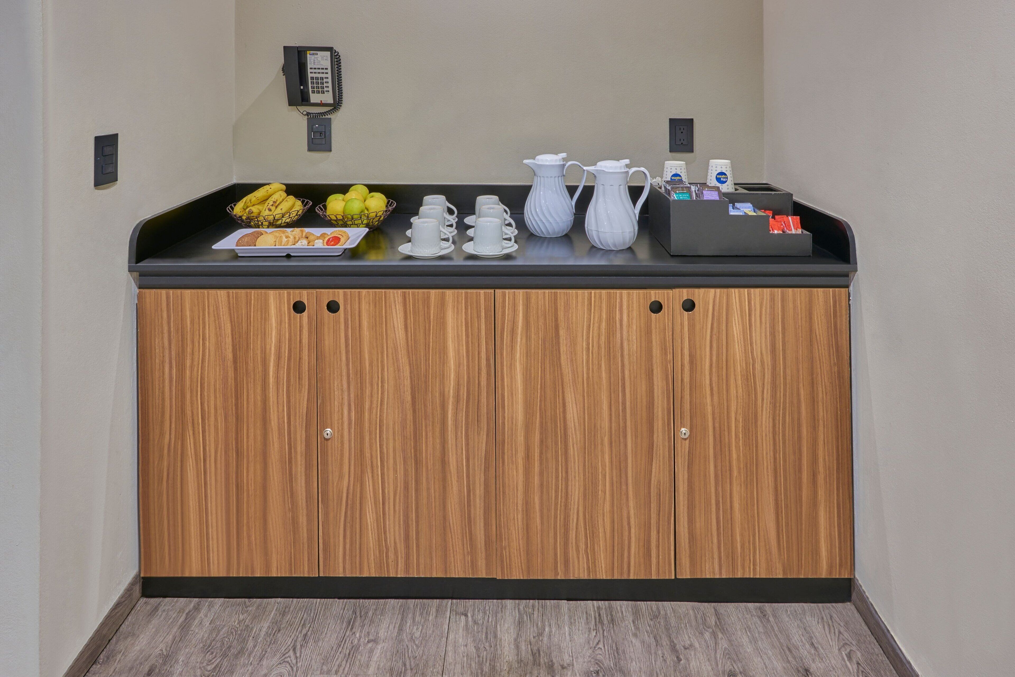 A kitchenette with a wooden cabinet, a fruit basket, white cups, a pitcher, and a coffee maker.