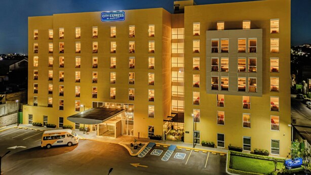 Un hotel de varios pisos con un llamativo letrero de 'City Express by Marriott', un área de estacionamiento techada y una vista despejada del paisaje urbano circundante.