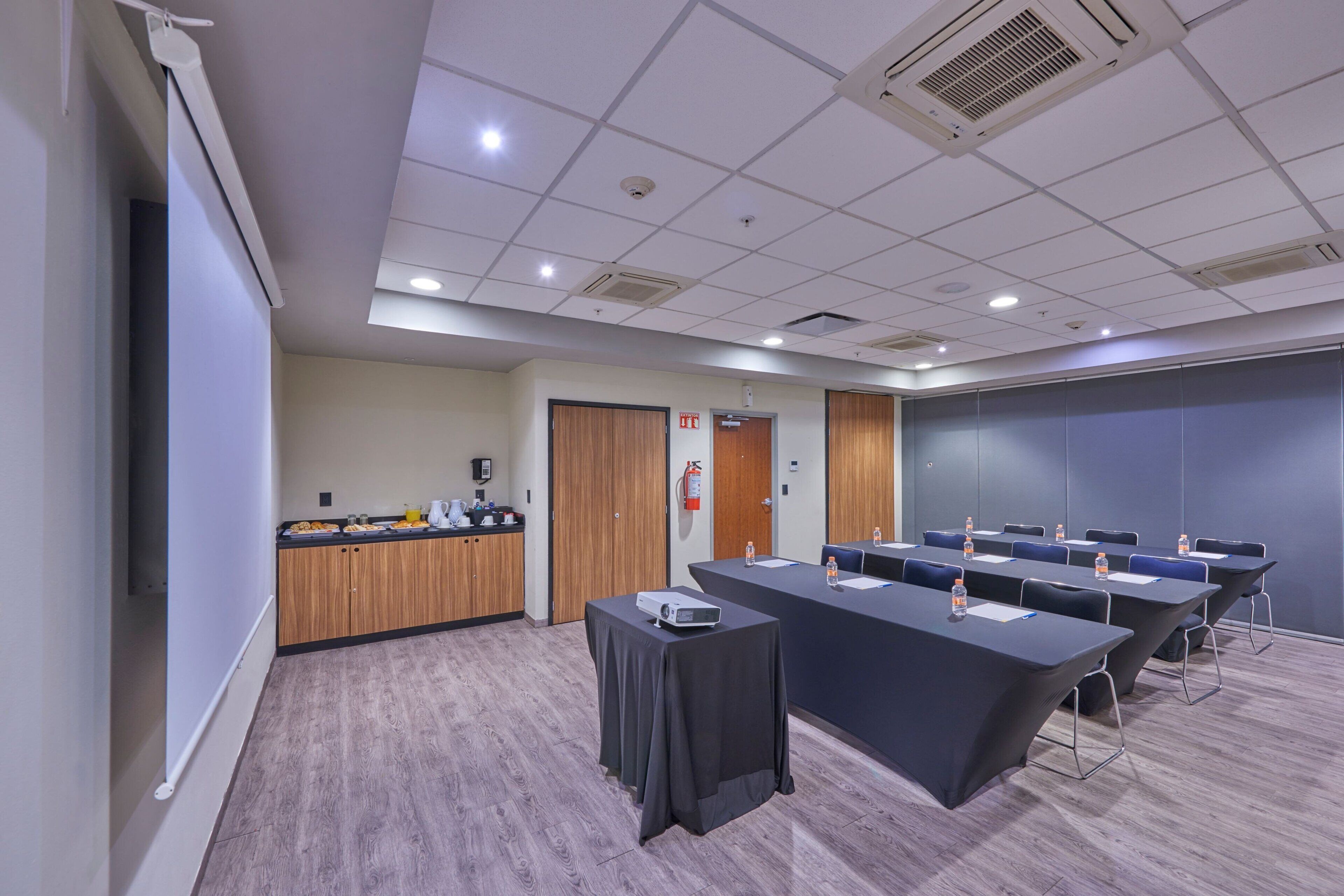 A conference room with a long table, chairs, a projector, and a small table with a water bottle.