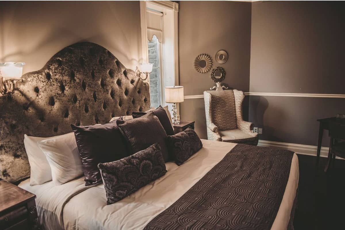 Un dormitorio con cabecera capitonada, colcha estampada y una silla con cojín.
