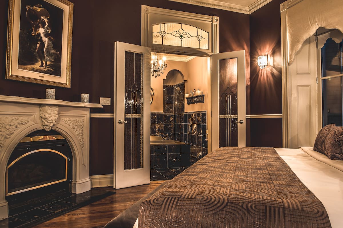 Un dormitorio con una cama grande, chimenea, candelabro y baño visible a través de una puerta abierta.