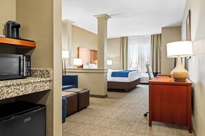 A hotel room with a kitchenette, a bed, a desk, and a lamp. - Heartland Inn and Suites (Wheatland)