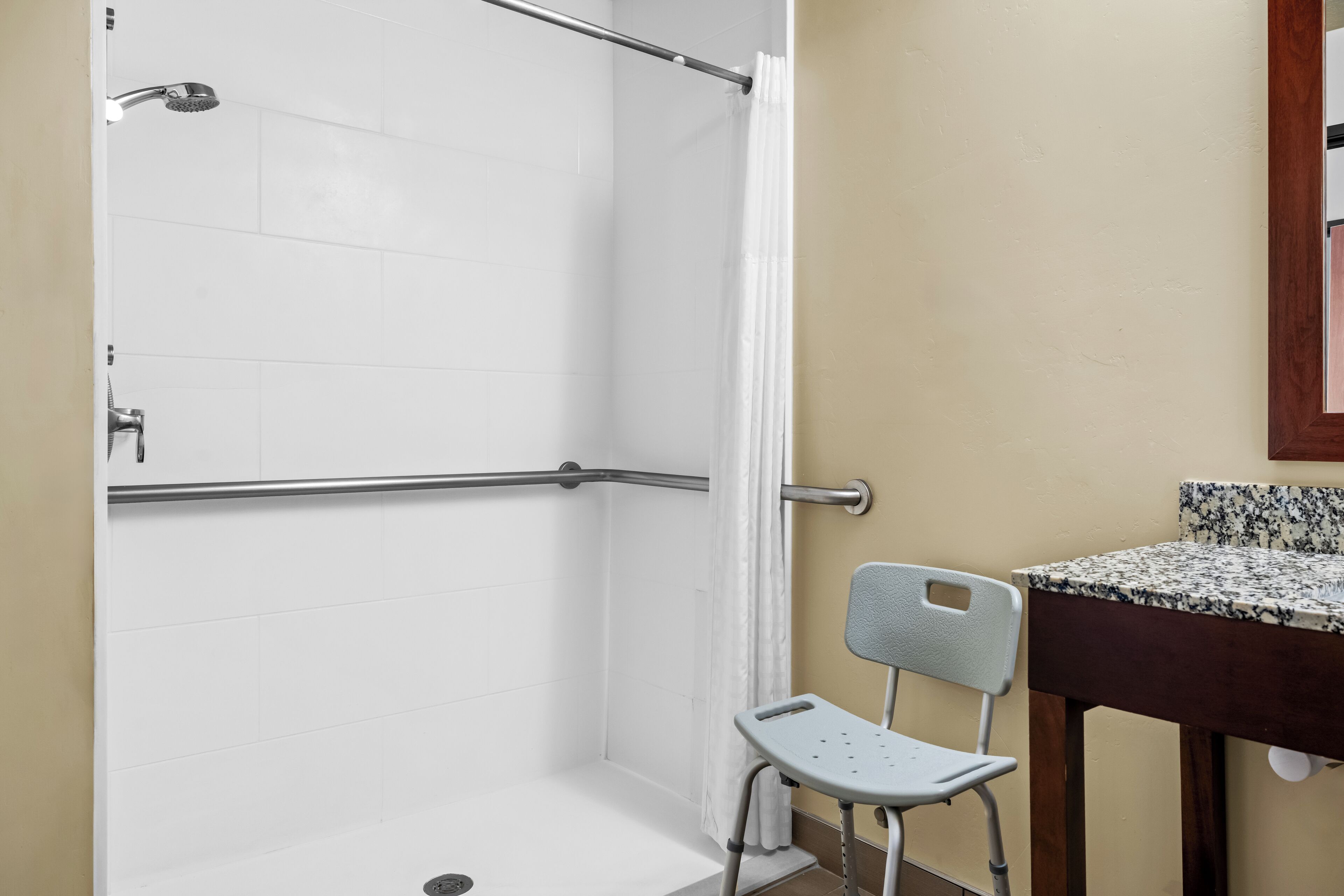 Zona de ducha con barra de apoyo, una silla y un lavabo con encimera de granito.