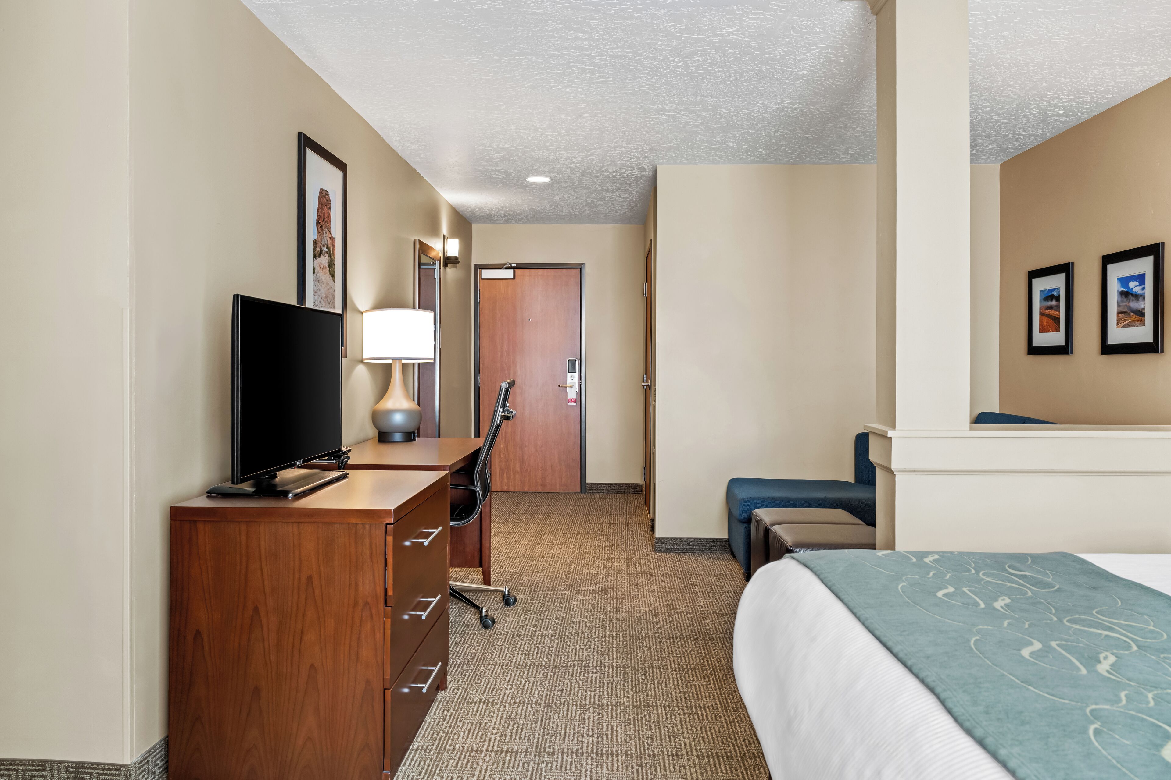 A hotel room with a wooden dresser, a flat-screen TV, a desk lamp, a bed with a blue blanket, and a door leading to another room.