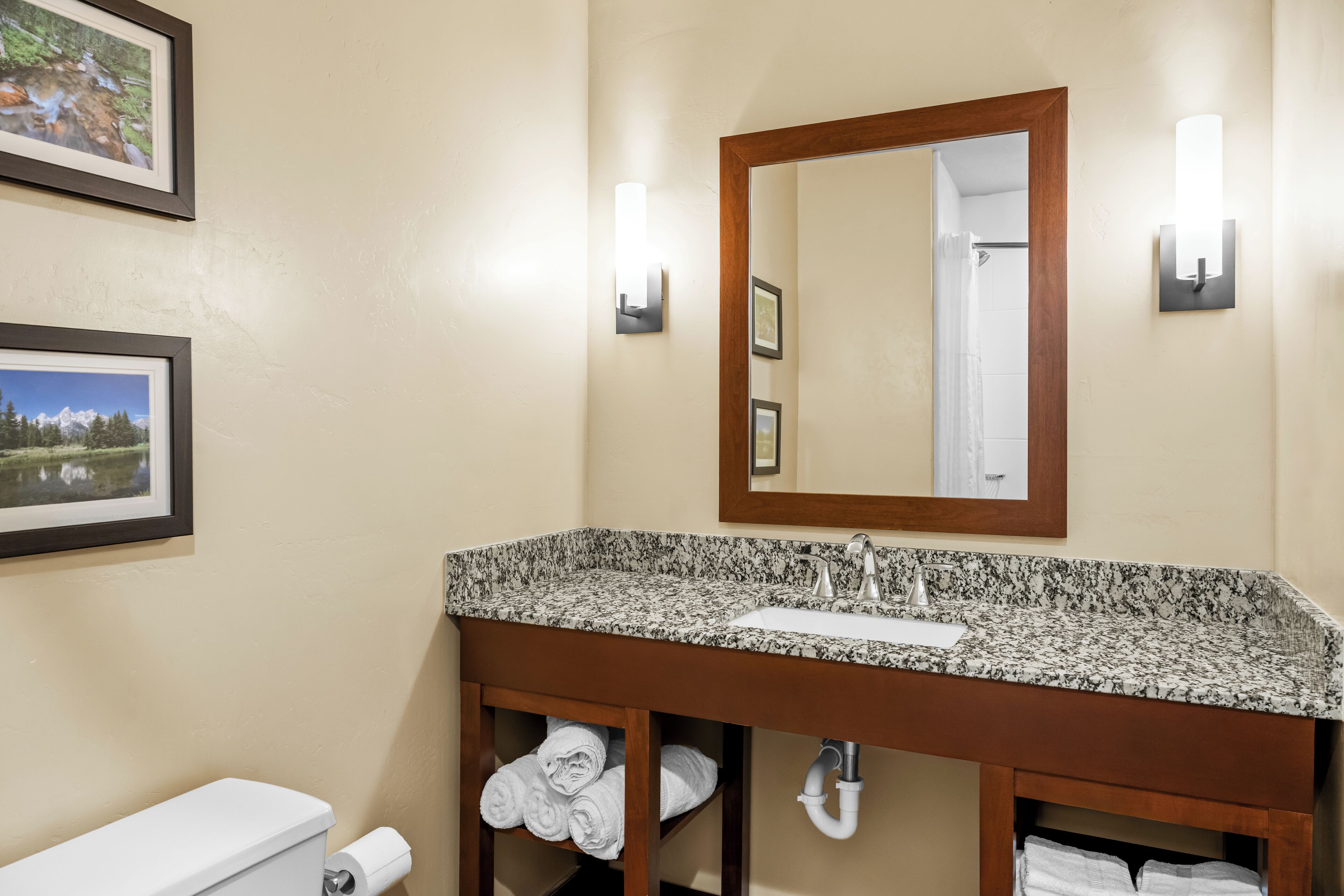 A bathroom with a granite countertop sink, a wooden vanity, a toilet, and two framed pictures on the wall.