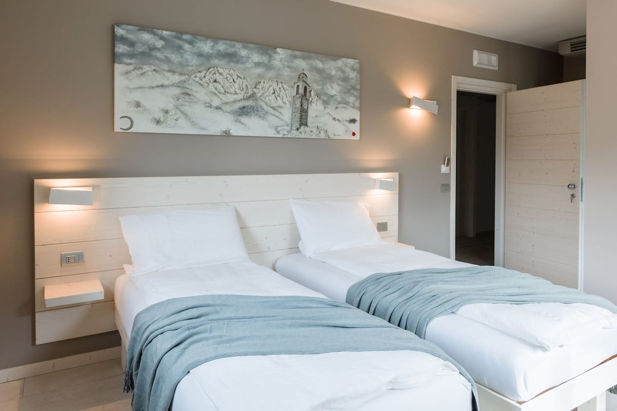Un lit double avec une literie blanche et un plaid bleu, une table de chevet avec une lampe, et un tableau représentant un paysage de montagne accroché au mur.