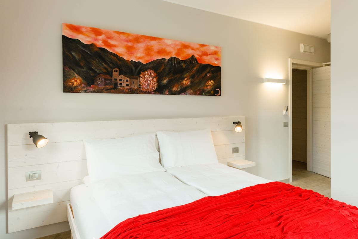 Une chambre à coucher avec un lit, une couverture rouge, un tableau représentant un paysage de montagne et une tête de lit blanche.