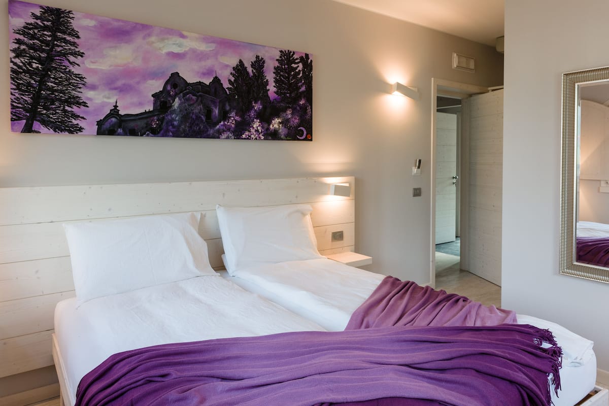 Une chambre à coucher avec un lit, du linge de lit blanc, une couverture violette, un tableau représentant des arbres et des montagnes, et un miroir accroché au mur.