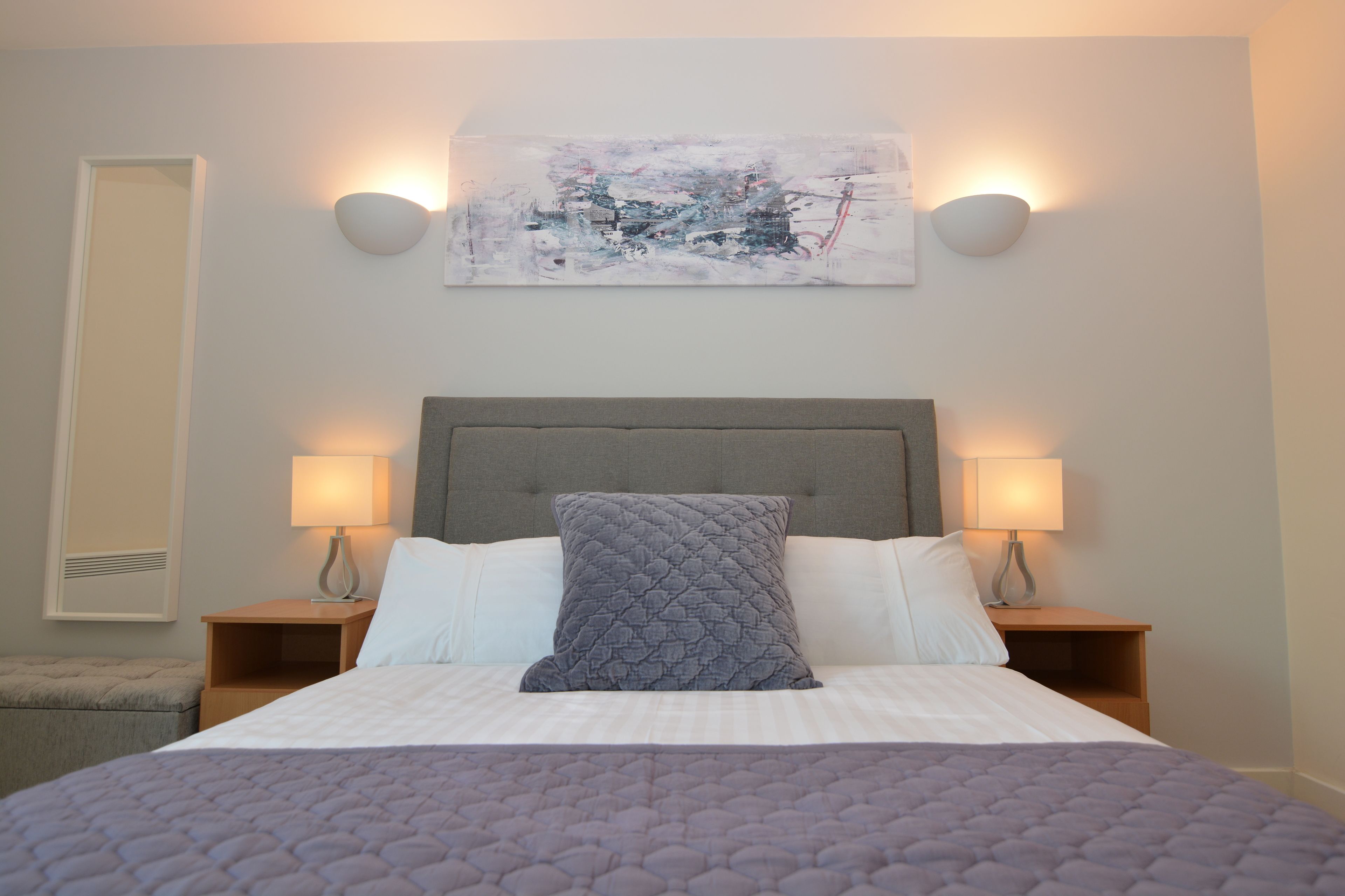 Un lit bien fait, recouvert d'une courtepointe matelassée, deux lampes de chevet et un tableau abstrait accroché au mur.