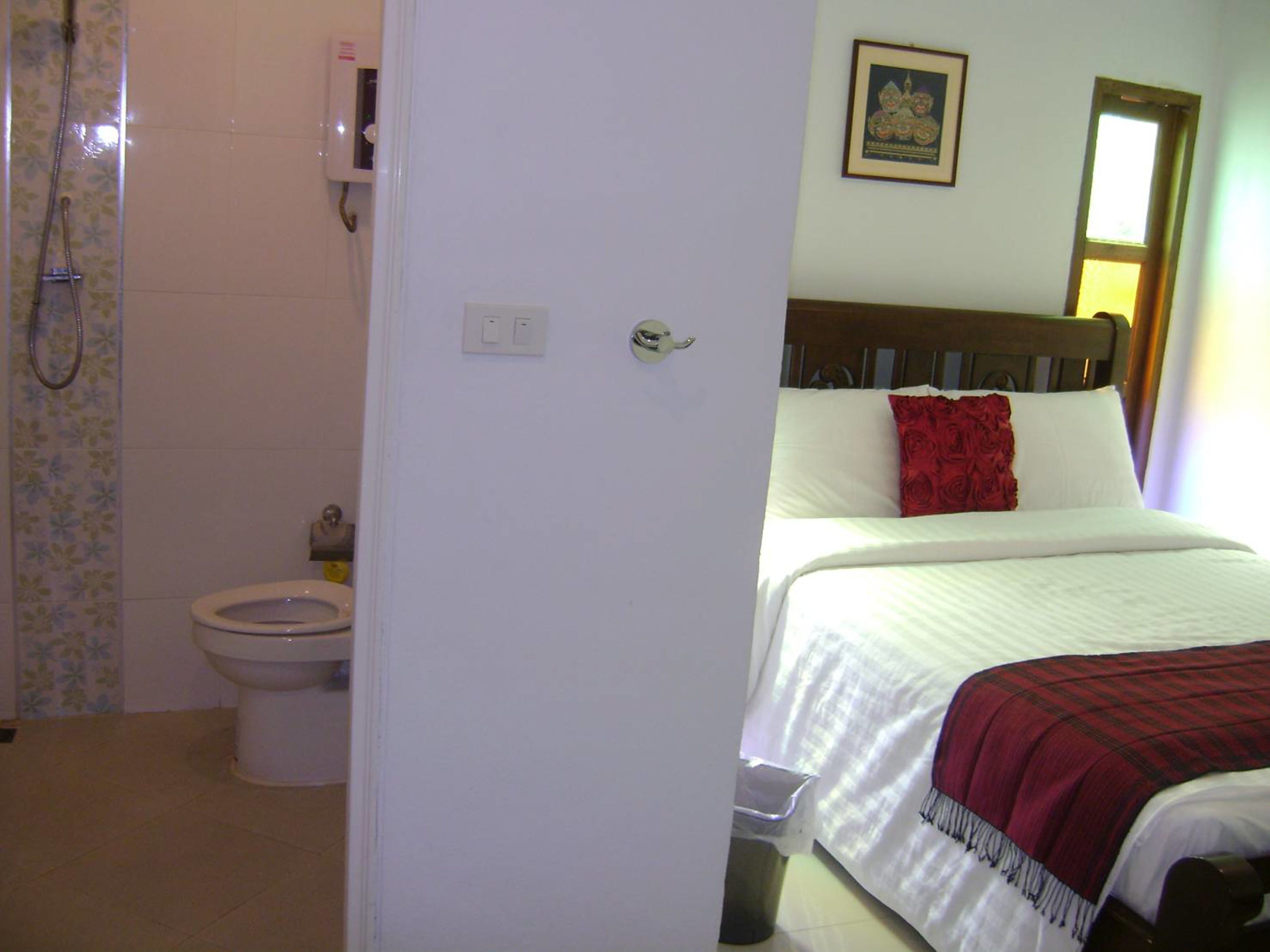 Une chambre à coucher comprenant un lit, une table de chevet, un oreiller rouge, une tête de lit en bois, une fenêtre et une salle de bain avec des toilettes et une douche.