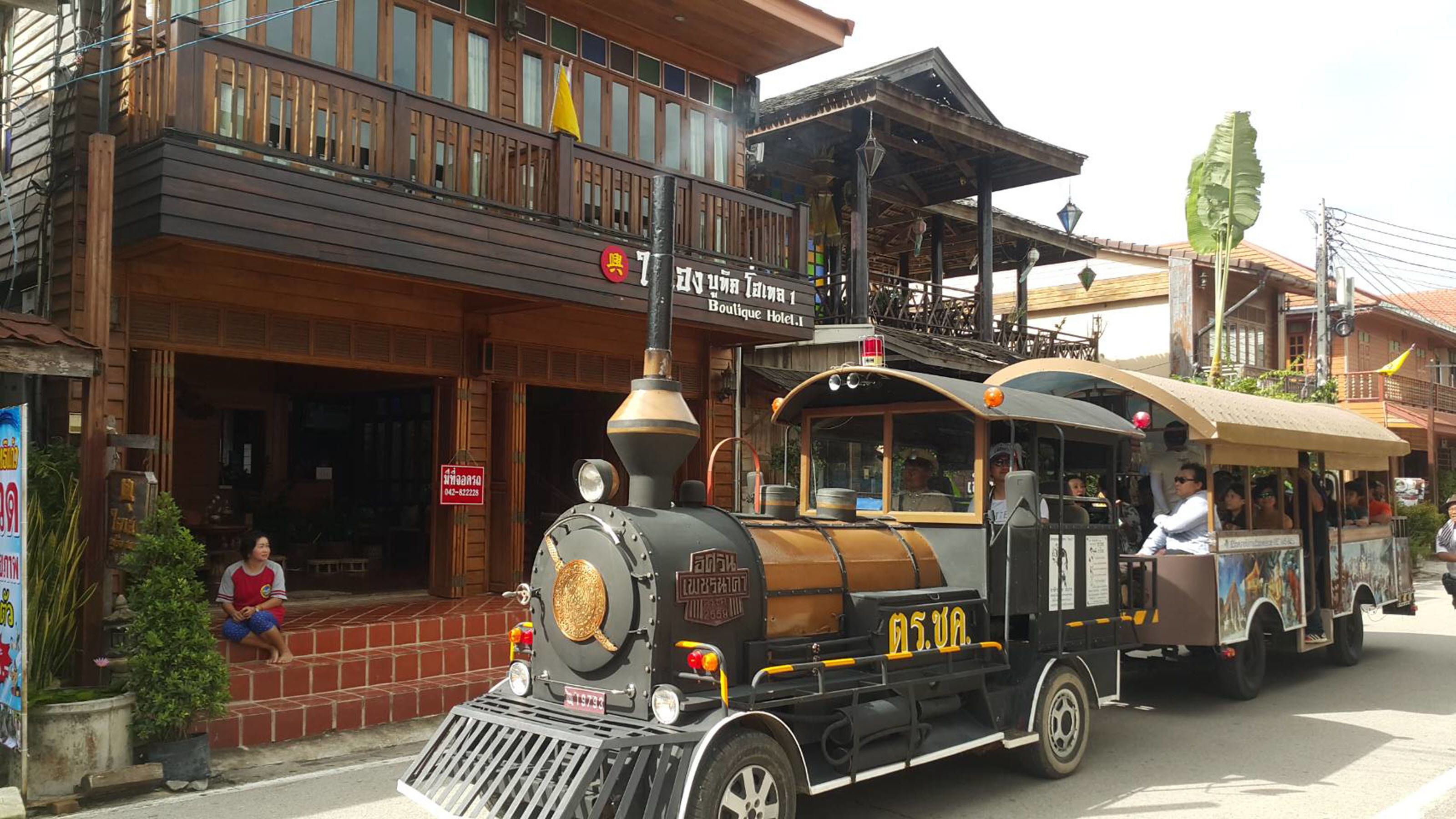 Un tren de vapor antiguo con pasajeros, pasando por un hotel boutique con balcones de madera y un cartel en un idioma extranjero.