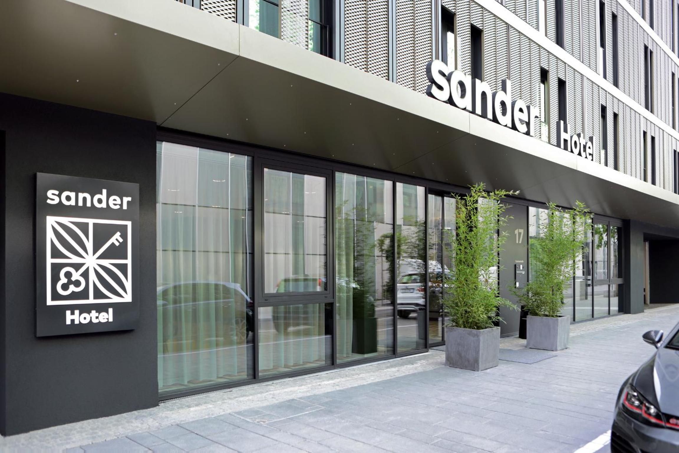 L’extérieur d’un hôtel, avec une façade moderne, de grandes fenêtres et une enseigne indiquant qu’il s’agit de l’hôtel « Sander ».