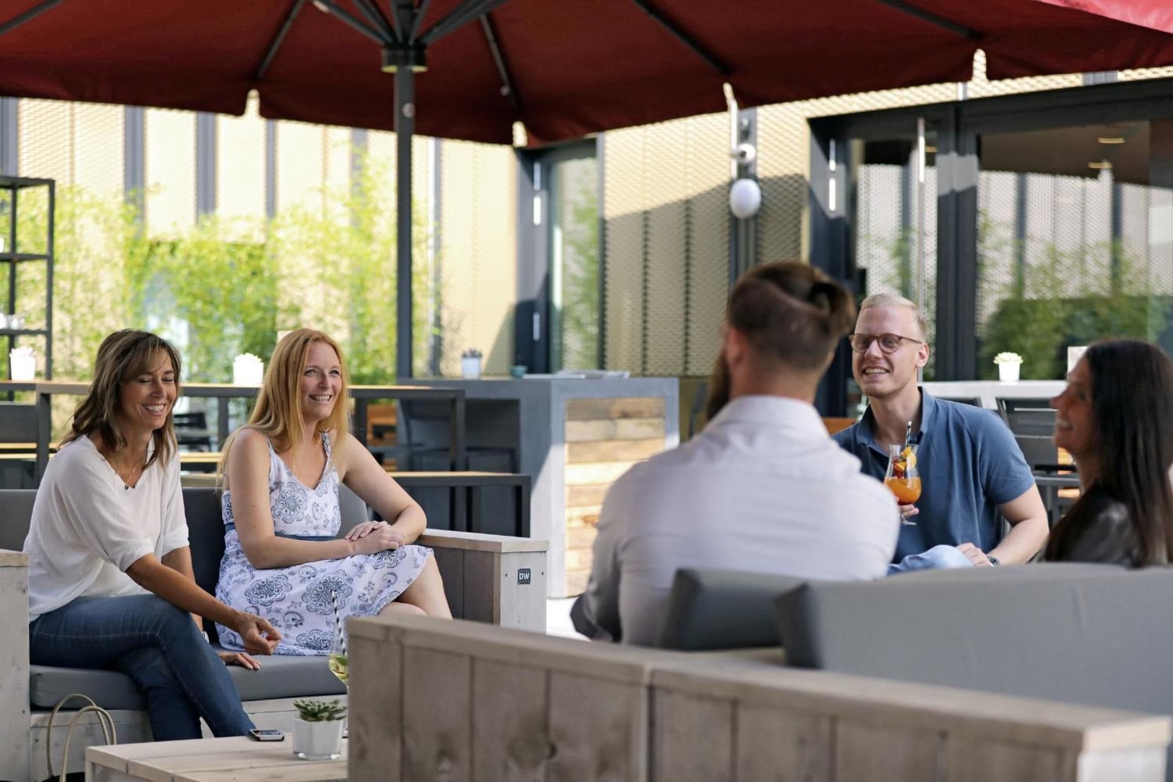 A group of people seated at an outdoor cafe with a red umbrella overhead.