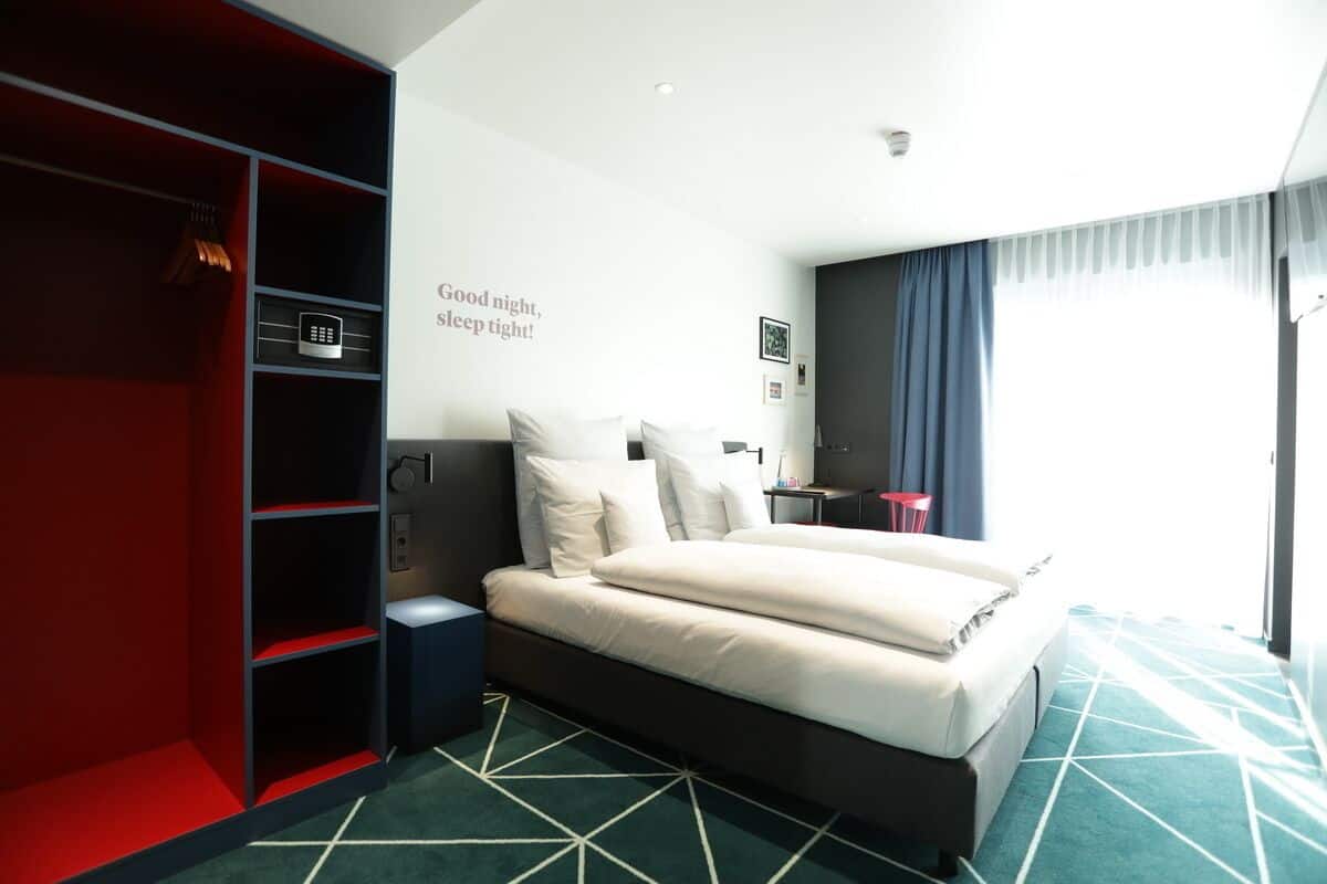 Una habitación de hotel moderna con una cama, un armario rojo, un escritorio y una ventana con cortinas.