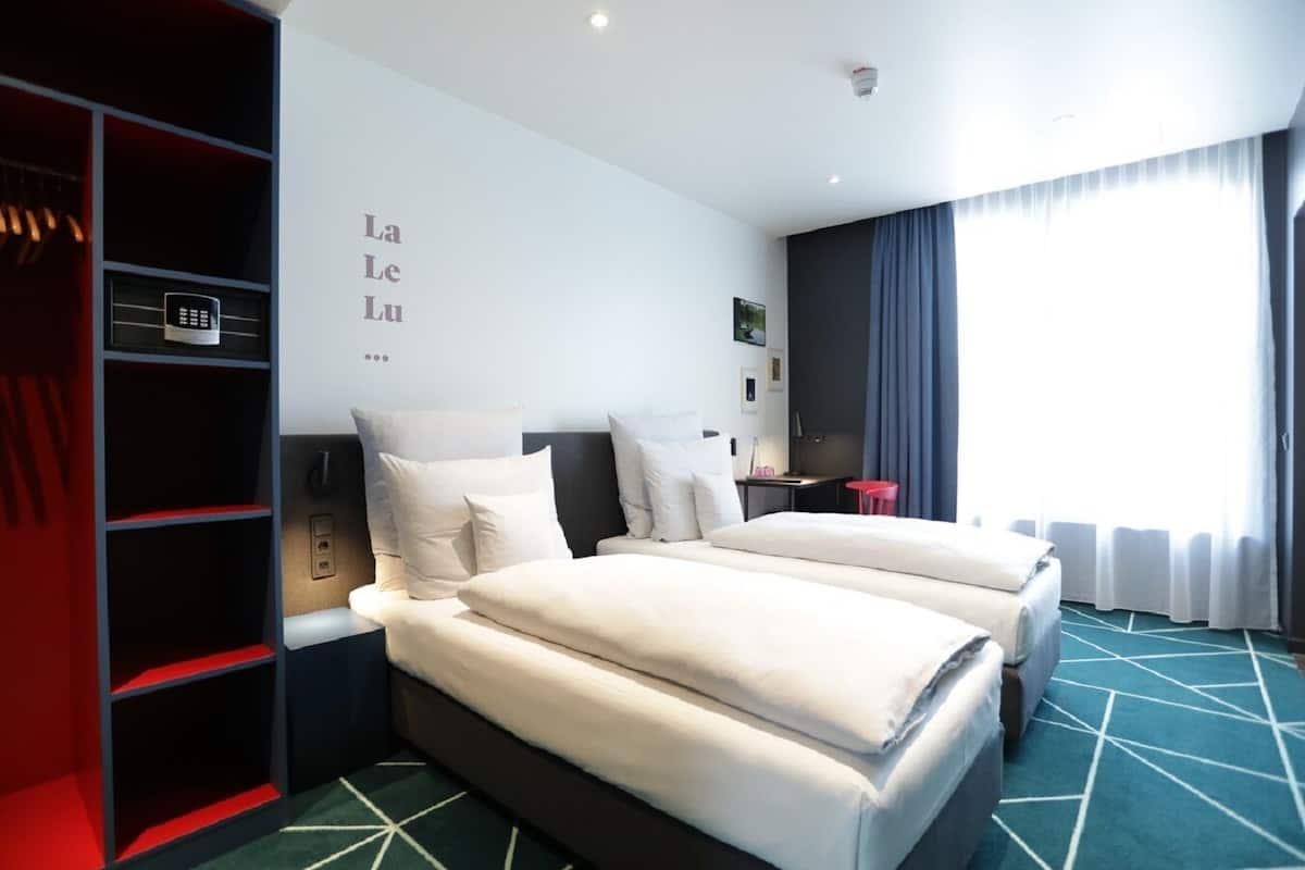 Una habitación de hotel con dos camas, un armario, un escritorio y una ventana con cortinas.