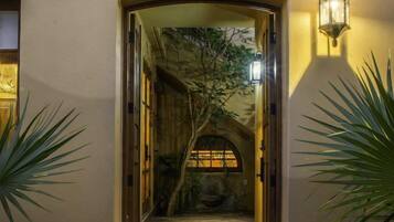 Uma porta em arco dá acesso a um alpendre coberto com chão de pedra, cercado por plantas em vasos e um candeeiro fixado na parede.