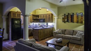 Un vaste espace de vie comprenant un canapé et une table basse, une cuisine avec des meubles en bois et un réfrigérateur, ainsi qu’un ventilateur de plafond.