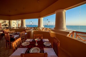 A dining area with tables set for two, large windows with a view of the sea, and a balcony with a railing.