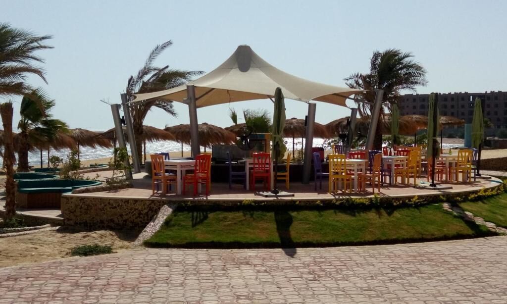 Ein Sitzbereich im Freien direkt am Strand mit Tischen und Stühlen unter einer Markise, umgeben von Palmen und einem Pool.