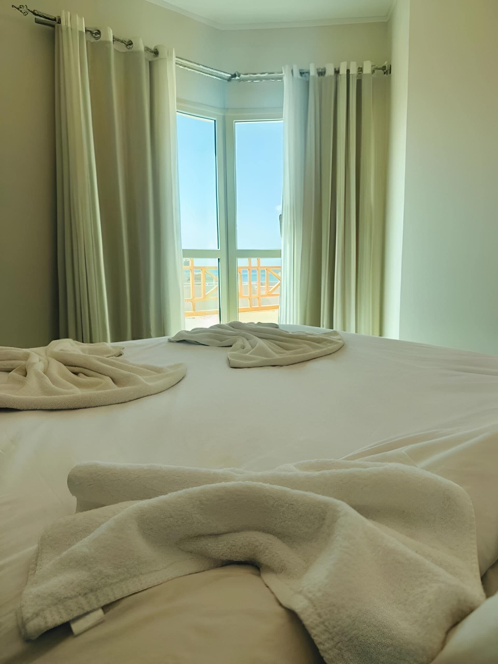 Habitación de hotel con cama, cortinas blancas y vistas a un balcón con barandilla.