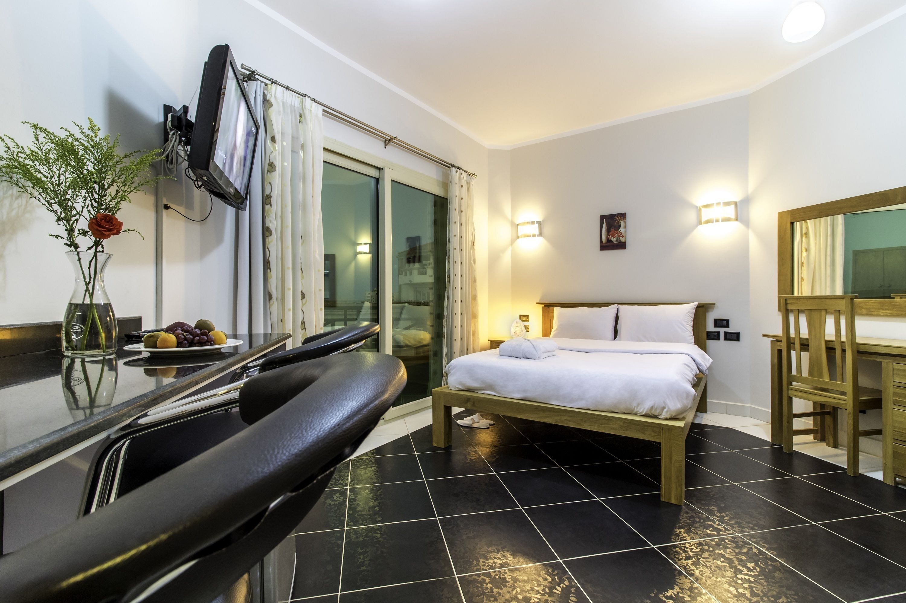 A hotel room with a bed, a desk, a chair, a small table with fruit, and a vase with a flower.
