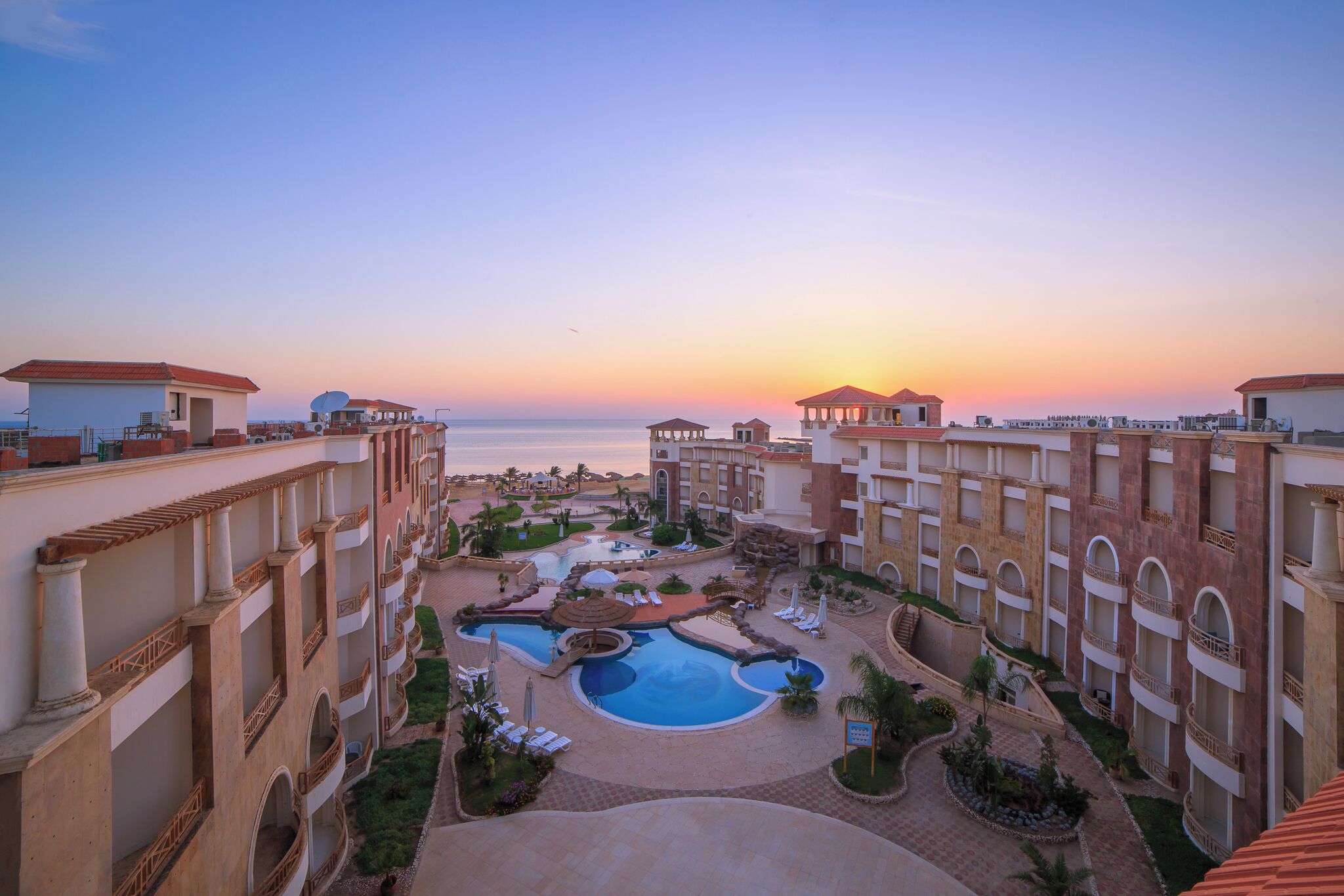 Un resort con varias piscinas, una fuente central y vista al océano al atardecer.