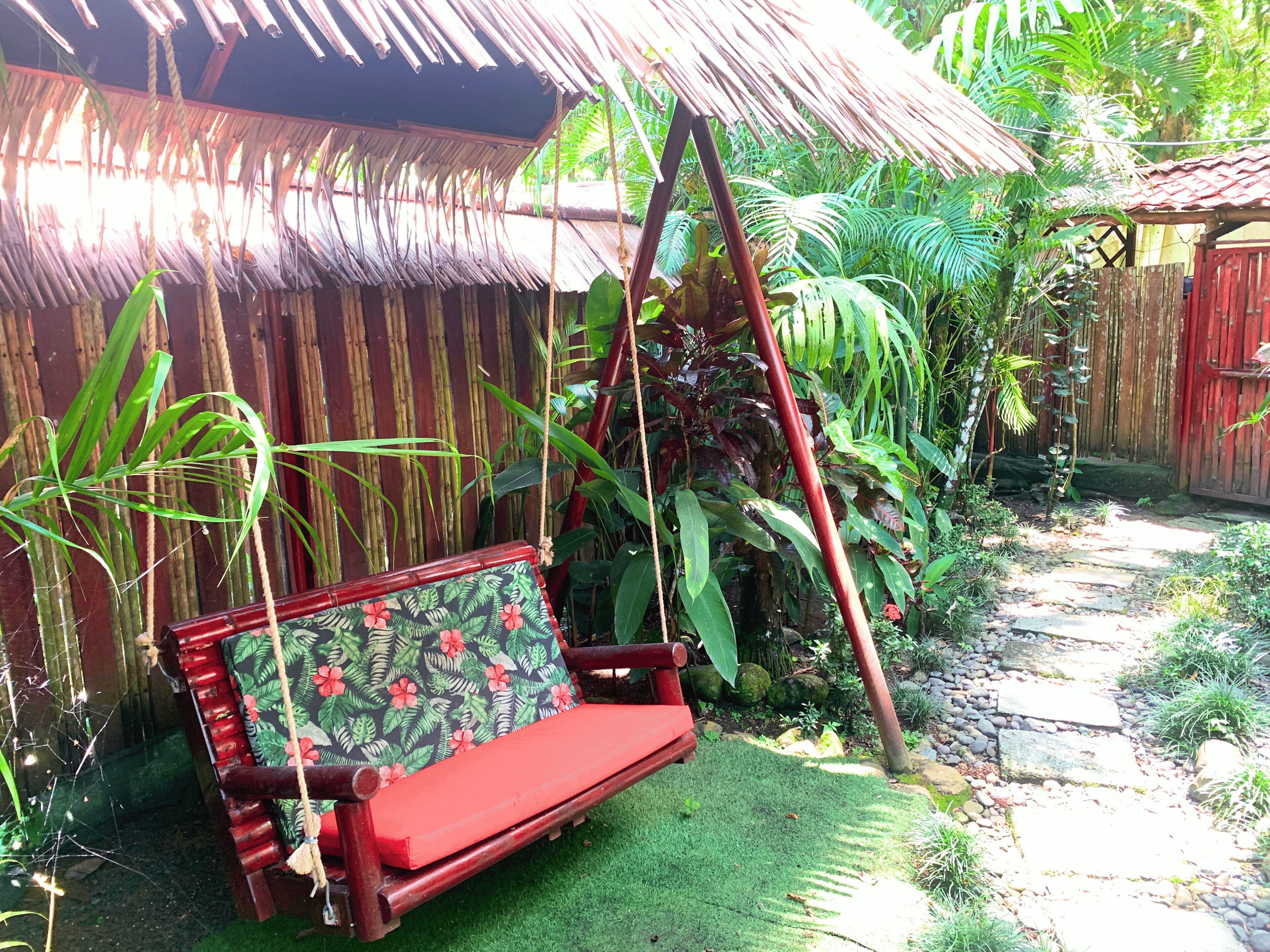 A wooden swing chair with a floral cushion in a garden with a thatched roof structure.
